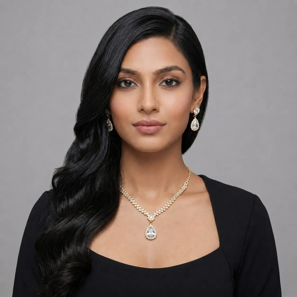 A woman with long, wavy black hair is shown from the chest up against a plain gray studio background. She is wearing a black top and a gold-tone necklace with a clear teardrop pendant, along with matching drop earrings.